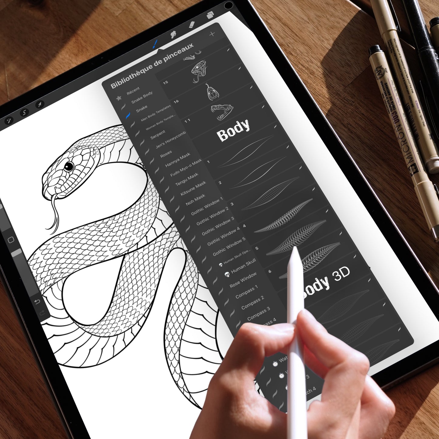 Hand holding an Apple Pencil using the Procreate app on an iPad, displaying a snake drawing created with Procreate brushes. The brush selection panel is open, highlighting the “Snake Body” and “Body 3D” brushes used to build detailed snake scale patterns. The iPad is placed on a wooden table with technical pens in the background.