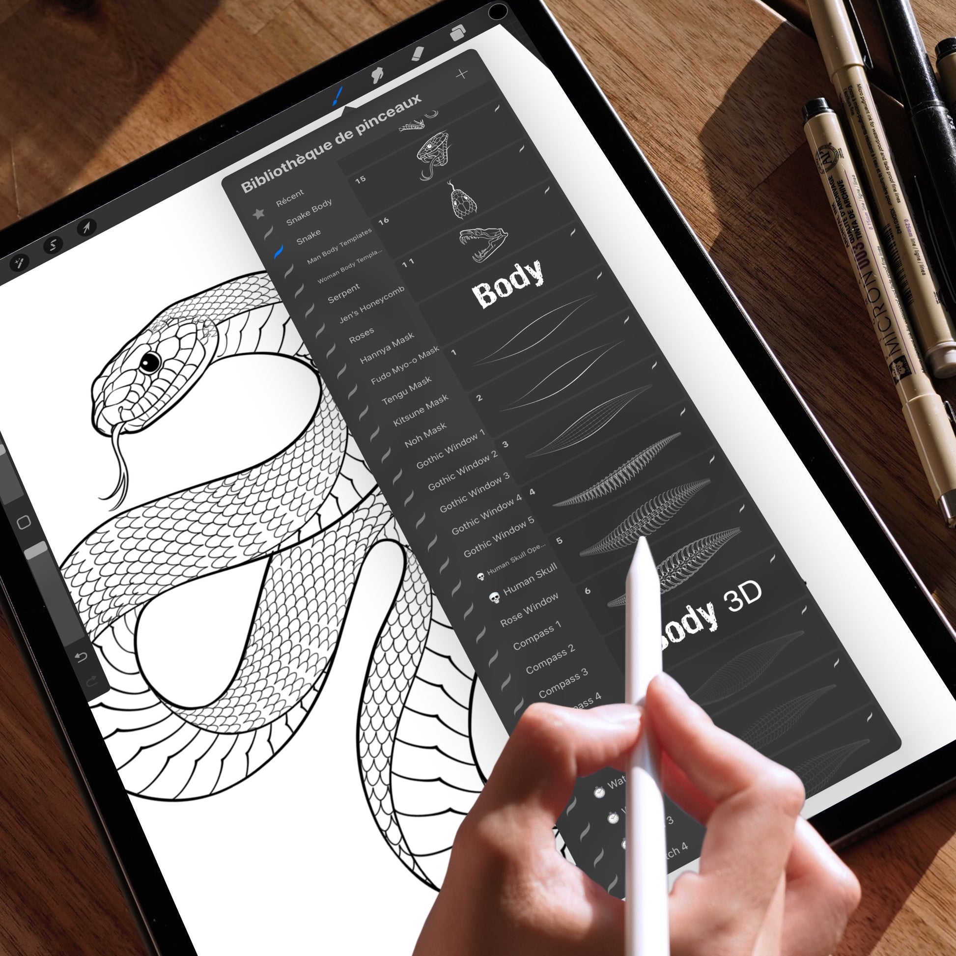 Hand holding an Apple Pencil using the Procreate app on an iPad, displaying a snake drawing created with Procreate brushes. The brush selection panel is open, highlighting the “Snake Body” and “Body 3D” brushes used to build detailed snake scale patterns. The iPad is placed on a wooden table with technical pens in the background.